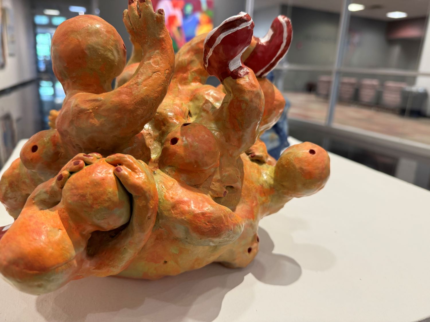 A colorful, abstract ceramic sculpture featuring a cluster of bulbous human figures, displayed on a white surface in a brightly lit room.