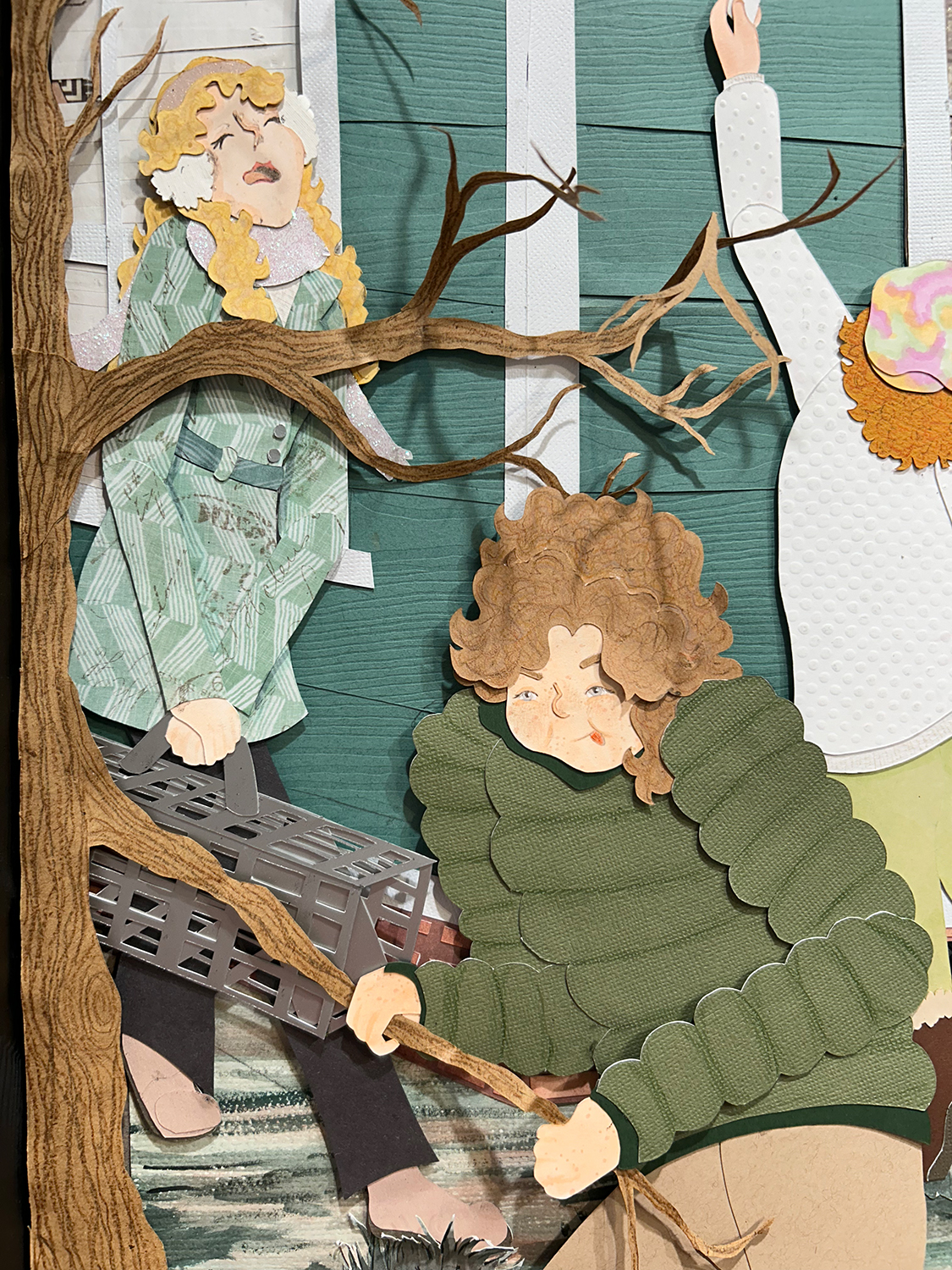 An image of a cut-paper scene. Three female figures are shown in front of the corner of a house. The figure on the left carries a silver animal cage. The figure in the middle foreground attempts to break off twig from a tree. The figure on the right reaches upward offering a marshmallow to a raccoon out of frame.