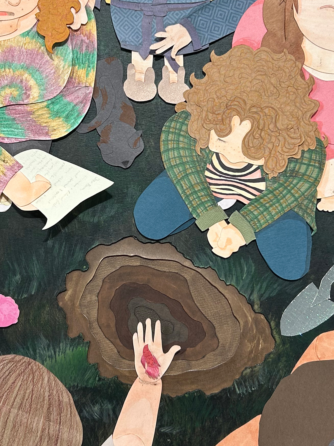 Image of a cut-paper scene. A figure crouches next to a hole in the ground. Above the hole another figure holds out her hand in the palm of which is a dead beta fish. Around the whole stand other mourners.