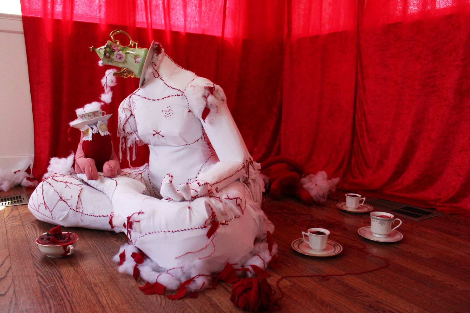 Figure of white fabric, red stitches, and a teapot for a head sits in a crossed legged position. A red fabric cat stands on the figure’s leg.  