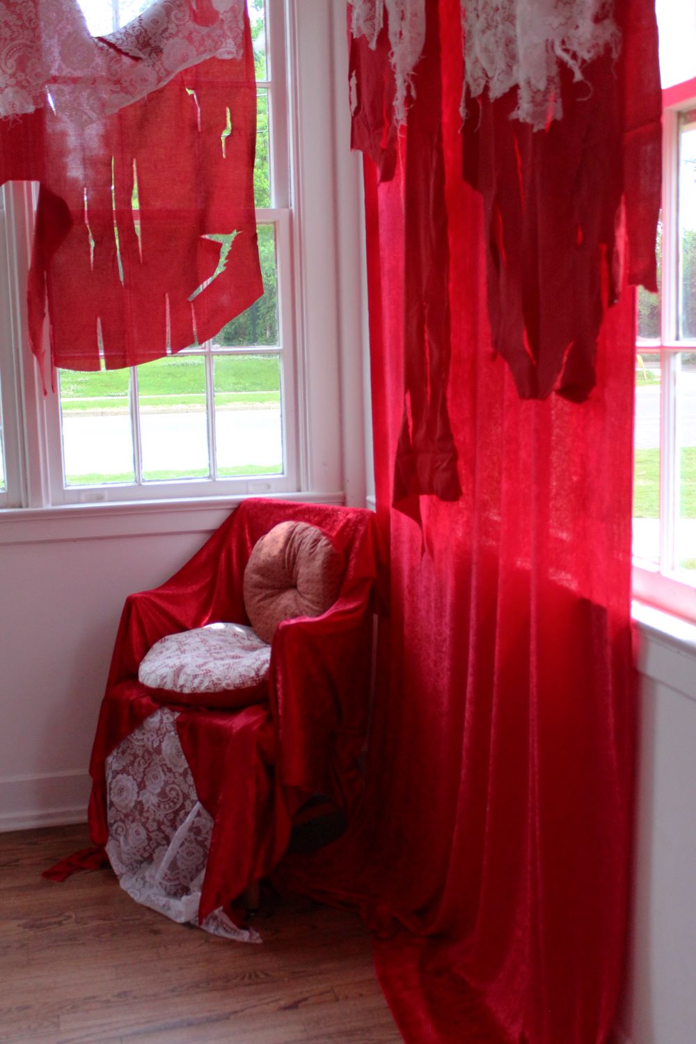 A cushioned chair draped in red, surrounded by tattered red curtains and white walls
